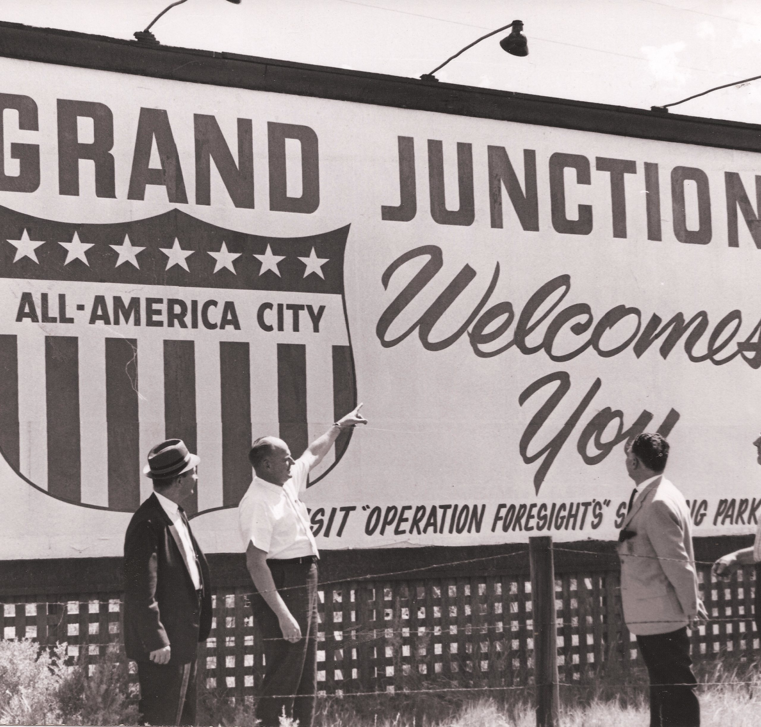 1963 black-and-white photo of several men looking at a "Grand Junction Welcomes You" billboard