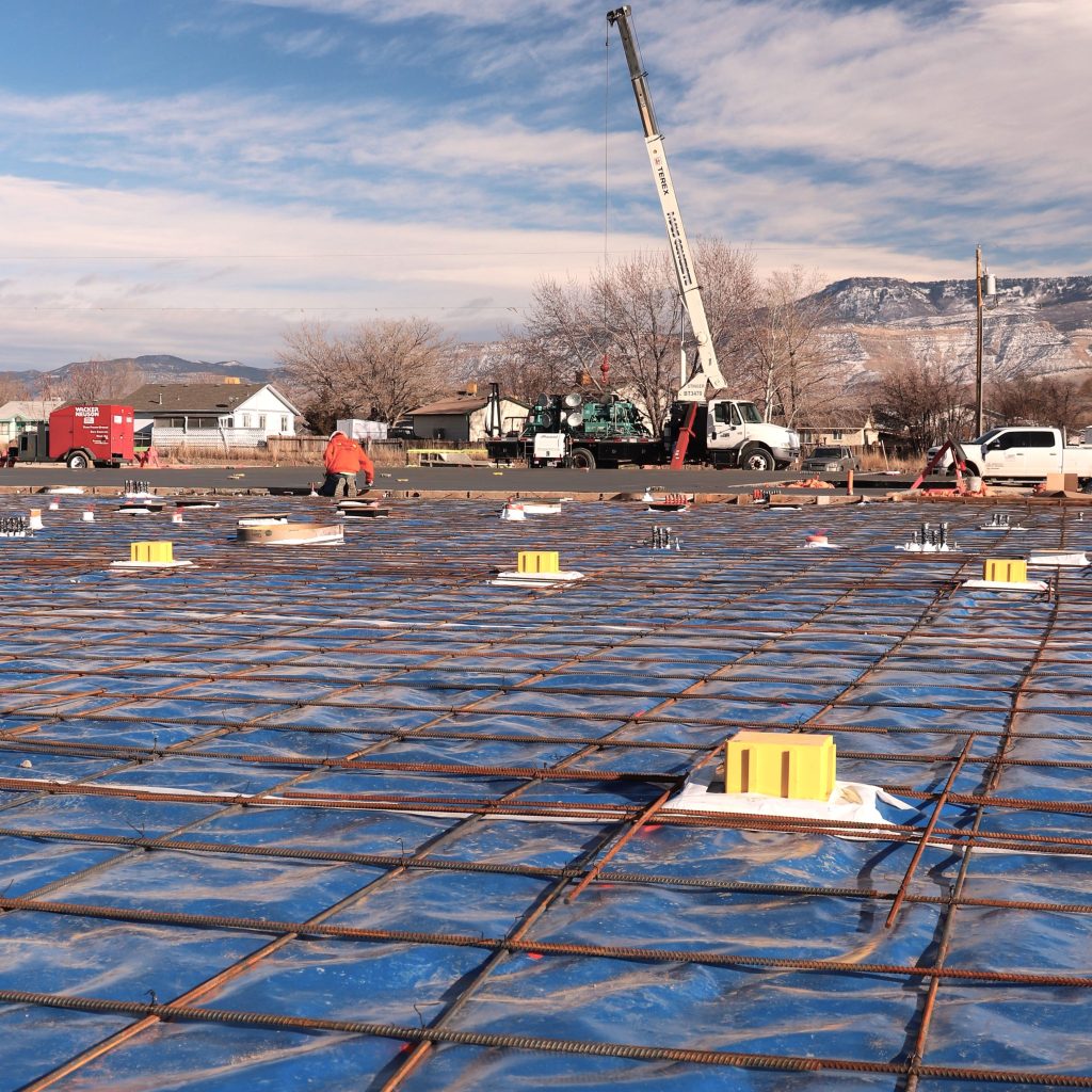 A grid of rebar sits atop a vapor barrier, with a portion of finished concrete foundation in the background.