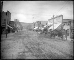 Photo I.D. - Black and white photograph depicting downtown Palisade, Colorado circa 1900-1908. It resembles a town from an old western movie, with dirt roads, horse-drawn carriages, and a saloon.