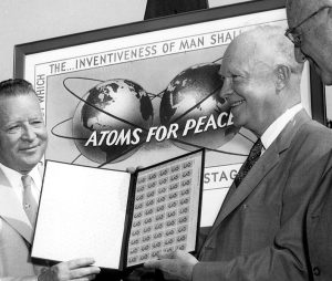 PHOTO I.D. - President Eisenhower displays an album with stamps promoting Atoms For Peace. The background includes a blown-up image of the stamp, depicting two globes with three rings, resembling an atom with electron rings. Partially cut off text reads "... To find the way by which the inventiveness of man shall be consecrated to his life," which was a quote from Eisenhower's Atoms for Peace speech.