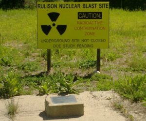 PHOTO I.D. - A shot of a yellow sign sitting behind an engraved plaque. The sign includes the trefoil ionizing radiation hazard symbol and reads "RULISON NUCLEAR BLAST SITE. CAUTION: RADIOACTIVE CONTAINMENT ZONE. UNDERGROUND SITE NOT CLOSED. DEPARTMENT OF ENERGY STUDY PENDING." The text on the plaque is not clearly visible.
