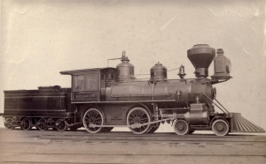 Photo I.D. - Sepia photograph depicting a Baldwin 8-18c Class narrow gauge locomotive with a 4-4-0 wheel arrangement. A single car is connected to the back of the engine. While not the same exact train as the Dread 107, both were produced by Baldwin Locomotive Works to meet the same specifications. 