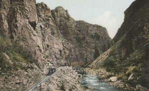 Postcard I.D. - Photo depicting a stretch of railway along the Black Canyon of the Gunnison River. A train can be seen along the rails in the bottom right corner of the image. The canyon cliffs tower over the scene, making the train appear very small against the natural landscape. 