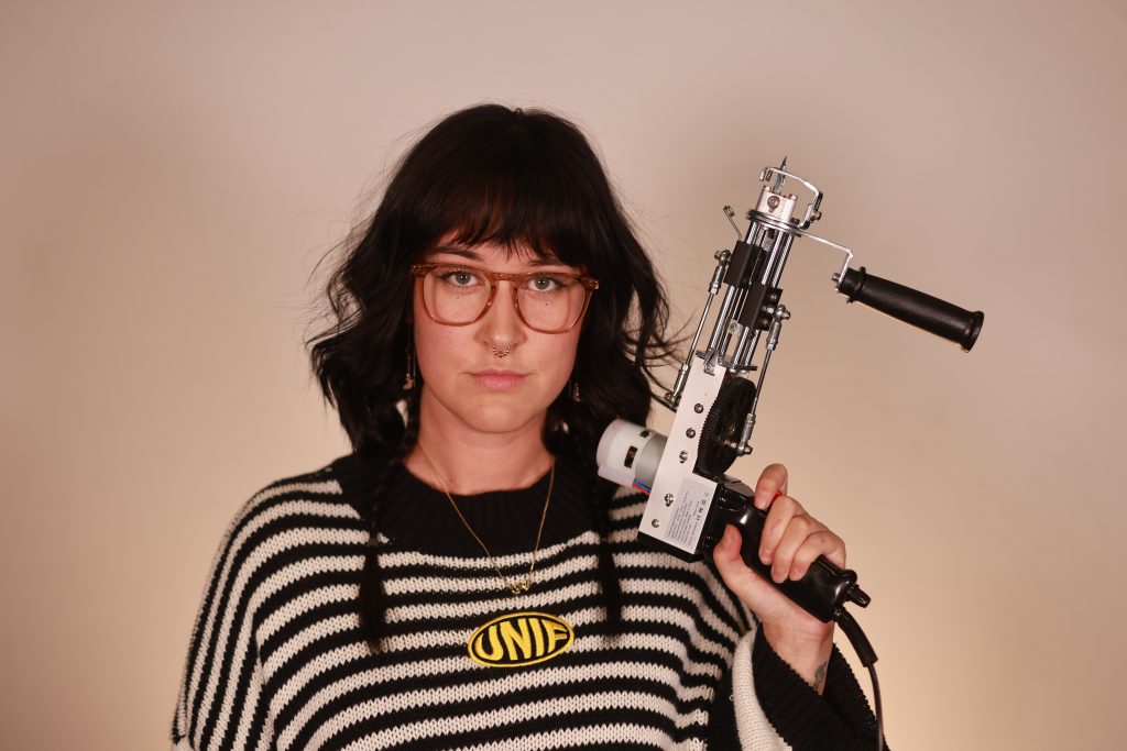 artist Emily Adamson wearing glasses, wearing a black and white striped shirt holding a rug tufting gun