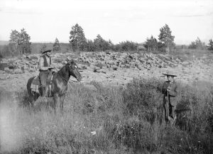 Photo depicting two sheep herders, an adult mounted on horseback and a child, on the La Sal Mountains in eastern Utah. A large herd of sheep can be seen grazing in the background.