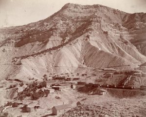 Sepia photograph depicting the town of Carpenter at the foot of the Book Cliffs. The town rests between two sandstone ridges. A train can be seen on the Little Book Cliff narrow gauge railway.