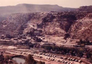 Color photo of the uranium and vanadium mill in Uravan, circa October 1952. Several cars and trailers are parked in a lot beneath the mill.