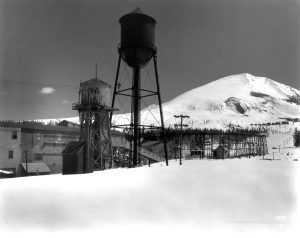 Black and white photograph depicting the crushing plant, water towers, and tramway at the Climax mine. There is a peak rising over the right side of the image. A thick blanket of snow covers the ground and the nearby peak.