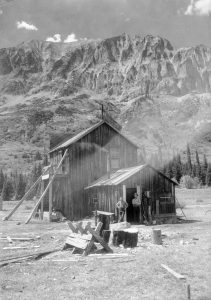 Black and white photograph depicting a weathered two-story wooden building that served as a saloon and a town hall. Two men are seen standing outside. Gothic Mountain towers over the building, with notable craggy pinnacles that are said to resemble gothic architecture.