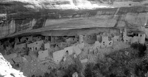 Black and white photograph of the Cliff Place in Mesa Verde National Park. The photo is dated to 1908, before any restoration efforts were undertaken. Several buildings are in ruin and there is debris scattered about in the walkways.