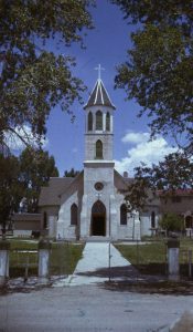 Color photograph of the Sangre de Cristo Catholic Church in San Luis. It has a central tower topped with a cross. 