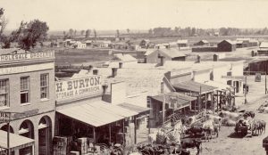 Black and white photo of Denver from the year 1860. Horse-drawn wagons and cows are parked on a dirt road in front of buildings labeled Wholesale Groceries," "H. Burton Storage & Commission," and "Missouri House." The hotel has other signs: "Bar Room," and "Boarding." The South Platte River is visible in the background.