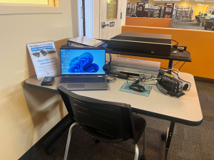 Table and black chair, tabletop has a binder, laptop, mouse, mousepad, vhs player, photo scanner, and camcorder.